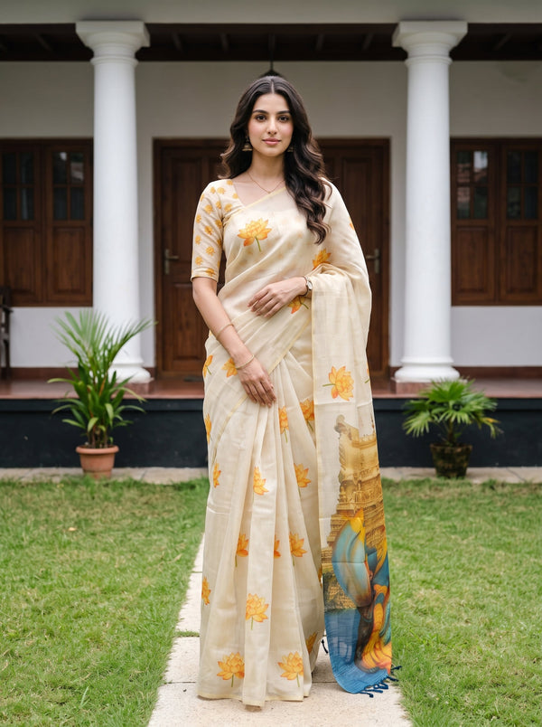A woman stands outdoors in front of a house with white columns and wooden doors, wearing a cream-colored saree with yellow floral motifs and a multicolored printed border. She is posed on a stone path with green grass and potted plants around her.