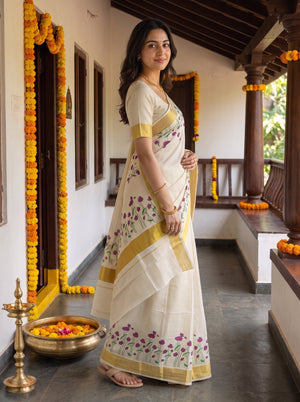 A woman wearing a cream-colored traditional Indian saree with yellow borders and purple floral embroidery, standing on a decorated outdoor porch.