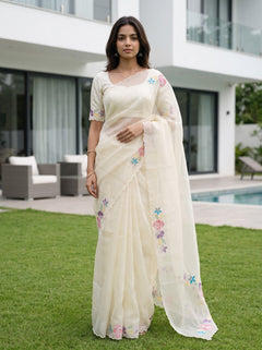 A woman stands on a green lawn in front of a modern house, wearing a cream-colored saree with pastel floral embroidery and a matching embroidered blouse.