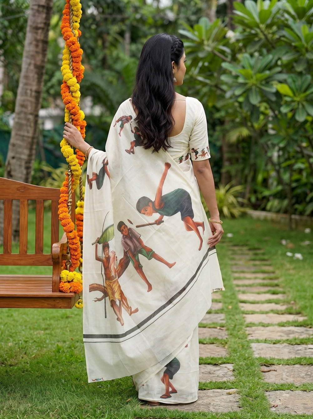 A woman wearing a cream-colored saree with a printed illustration of children is standing outdoors next to a wooden swing decorated with orange and yellow marigold garlands.