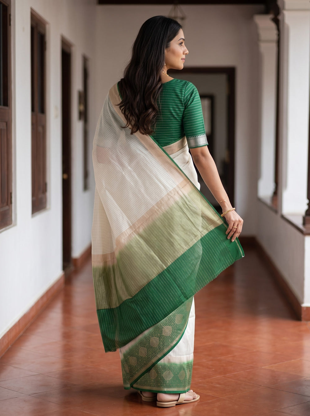 A woman wearing a white and green saree stands in a hallway with white walls and dark door frames, holding onto a wall for support.