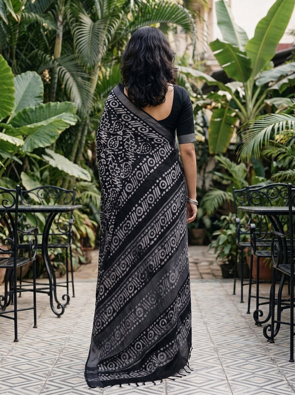 A woman viewed from behind wearing a black and white patterned saree in an outdoor garden patio setting with potted plants and black metal tables.
