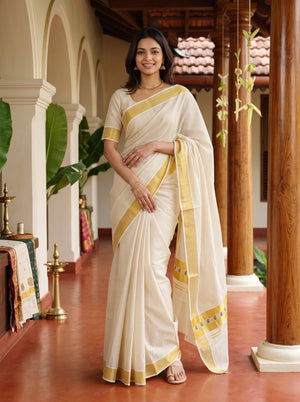 A woman wearing a cream-colored cotton saree with yellow-gold borders and a matching blouse, standing in a traditional corridor with wooden pillars and arches.