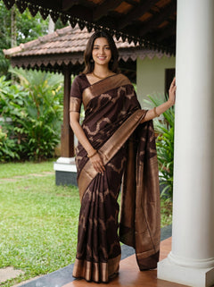 Woman in a brown saree standing outdoors with greenery in the background