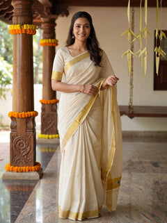 A woman wearing a cream-colored cotton saree with a yellow border stands in a traditional indoor setting with carved wooden pillars decorated with orange marigold garlands.