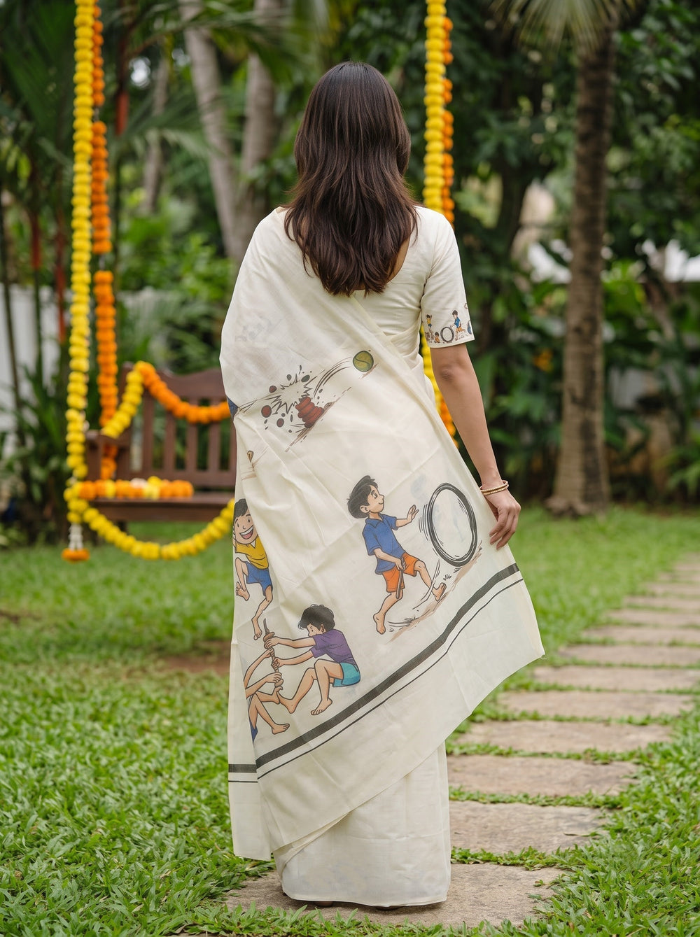 A woman viewed from behind wearing a cream-colored cotton saree with colorful traditional Indian folk art illustrations, standing on a stone path in a garden.