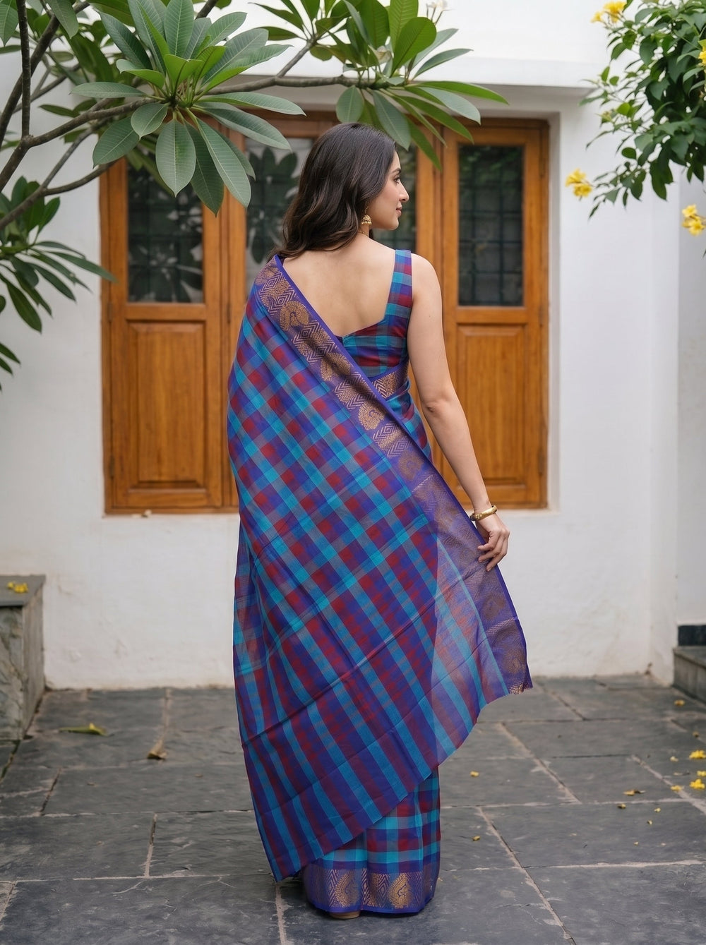A woman wearing a purple and blue plaid silk saree with gold zari detailing, standing outdoors in front of a white wall with wooden doors.