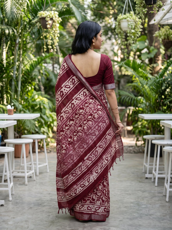 A woman wearing a deep maroon saree with beige floral patterns, photographed from behind in an outdoor garden setting with white tables and stools.