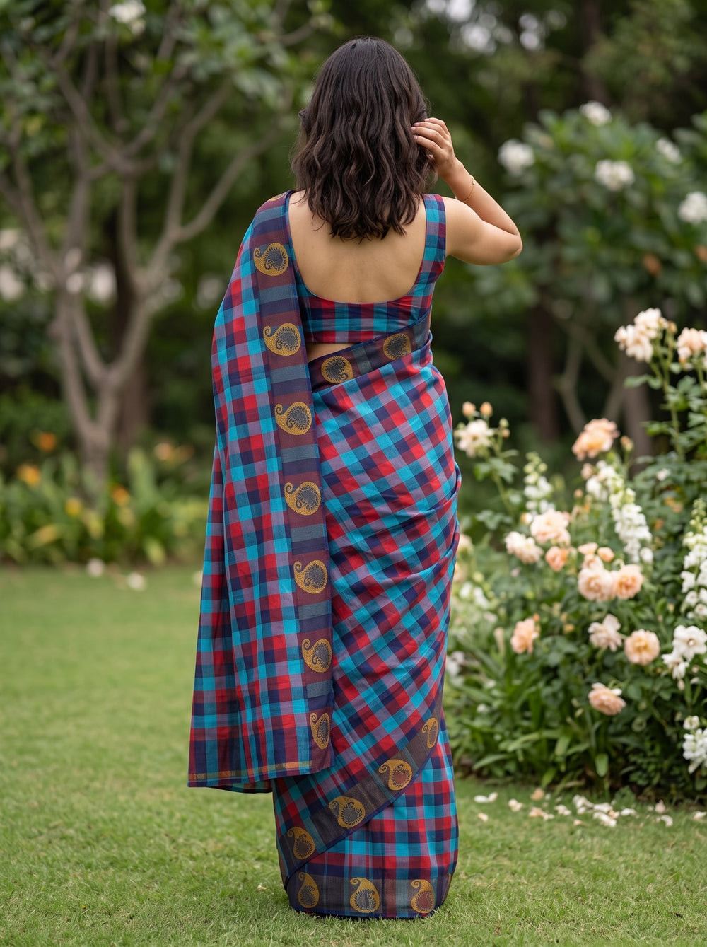 A woman wearing a blue and red plaid saree with gold motifs stands outdoors on a grassy lawn with trees in the background.