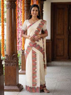 A woman wearing a cream-colored cotton saree with a red, orange, and green geometric patterned border, standing in a traditional indoor setting with wooden pillars and garlands.