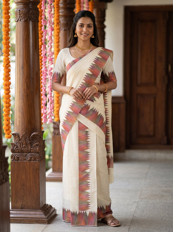 A woman wearing a cream-colored cotton saree with a red, orange, and green geometric patterned border, standing in a traditional indoor setting with wooden pillars and garlands.