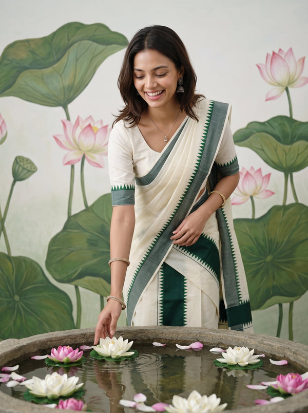 A woman wearing a cream and dark green saree stands beside a stone basin filled with lotus flowers, with a painted lotus mural in the background.