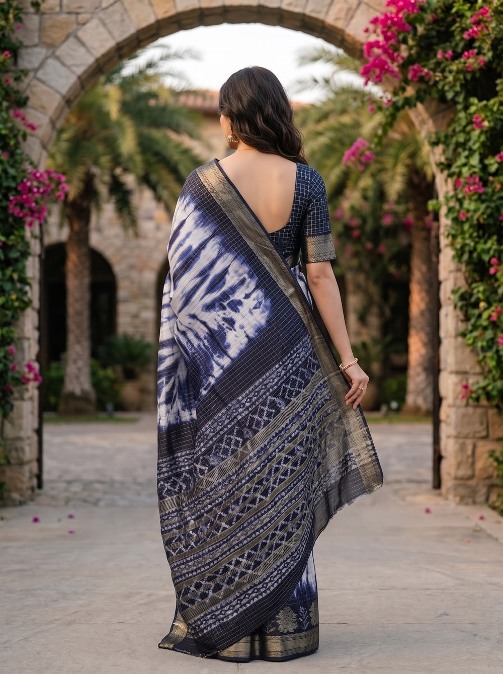 A woman models a blue and white tie-dye saree with a metallic gold border, standing in front of a stone archway decorated with greenery and pink flowers.