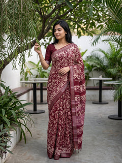 A woman wearing a deep maroon saree with a cream floral block print pattern, standing in an outdoor patio setting with green plants and tables.