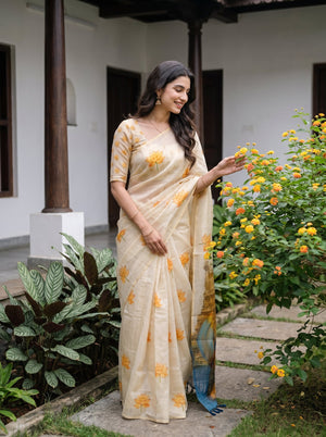 A woman wearing a cream-colored saree with yellow floral prints and a blue border, standing in a garden courtyard next to yellow flowers.
