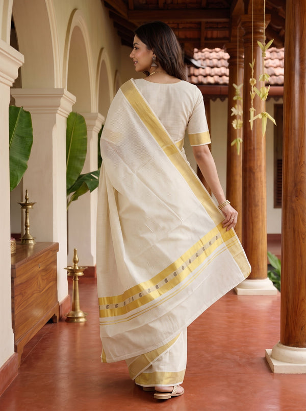 A woman wearing a cream-colored cotton saree with a gold border and a matching cream blouse, standing in a traditional courtyard with wooden pillars.