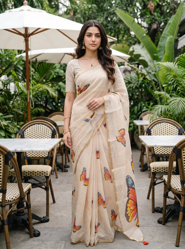 A woman wearing a beige saree with colorful butterfly prints stands in an outdoor patio setting with tables and umbrellas.