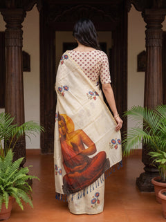 A woman is shown from behind wearing a cream-colored saree with a printed illustration of a seated figure and blue floral motifs, standing in a traditional courtyard with dark wooden pillars and potted plants.