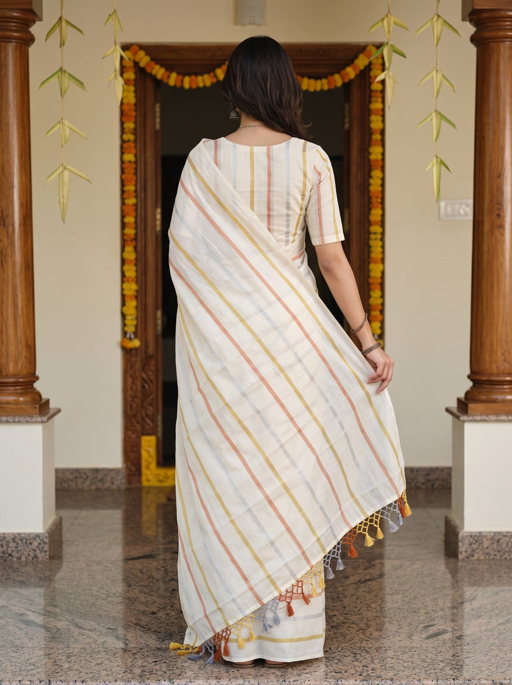 A woman wearing a white cotton saree with thin orange and yellow stripes, standing in a decorated doorway with wooden pillars and marigold garlands.