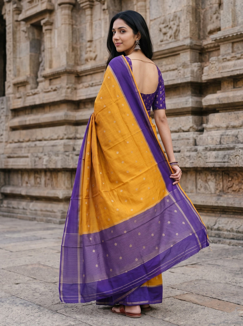 A woman wearing a yellow and purple silk saree stands in front of a stone temple structure. The saree has a yellow body with a purple border and a matching purple short-sleeve blouse.