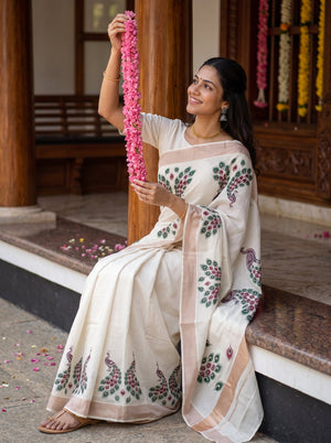 A woman wearing a cream-colored cotton saree with green and pink floral prints sits on stone steps in front of a wooden building, holding a pink flower garland.