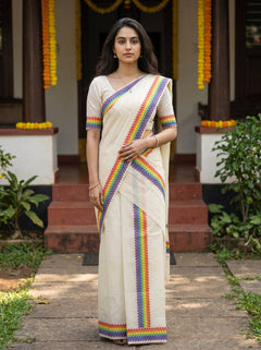 A woman stands outdoors in front of a doorway wearing a cream-colored sari with vibrant multicolored striped borders. The sari has short sleeves with matching striped cuffs, and she is wearing a simple white blouse.