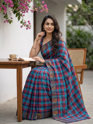 A woman wearing a blue and red plaid saree with gold motifs sits at a wooden table in an outdoor patio setting.