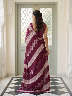 A woman stands with her back to the camera wearing a deep maroon saree with white block prints and a matching printed blouse, posed in front of a light green door in an elegant interior.
