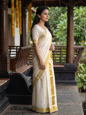 A woman wearing a cream-colored cotton saree with gold zari borders and a matching cream blouse, standing in a traditional wooden porch with green foliage in the background.