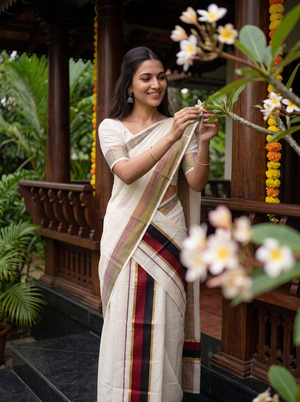 A woman wearing a traditional cream-colored saree with red, green, and gold stripes stands in a garden pavilion, holding white flowers.