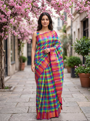 A woman stands outdoors on a stone walkway under pink flowering trees, wearing a bright multicolor plaid saree with a pink border and green, blue, yellow, and orange check pattern.