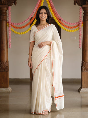 A woman wearing a cream-colored cotton saree with orange border stripes, standing in a decorated indoor space with wooden pillars and colorful garlands.