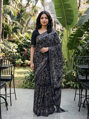 A woman wearing a black and white patterned saree with a black blouse, standing in a garden patio setting with plants and chairs in the background.