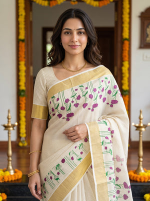 A woman wearing a cream-colored saree with purple floral patterns and green stems, paired with a matching cream blouse featuring gold borders. She is standing in a decorated indoor setting with brass lamps and orange garlands.
