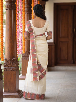 A woman viewed from behind wearing a cream-colored cotton saree with a red and green geometric patterned border and pallu, standing in a traditional courtyard with wooden pillars and hanging flower garlands.