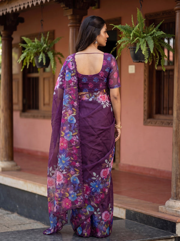 A woman wearing a purple floral printed saree with a matching blouse, standing on a porch with a pink wall and hanging green plants in the background.