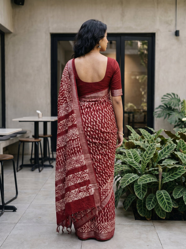 A woman wearing a deep red and beige patterned saree, viewed from behind as she stands in an outdoor patio setting with plants and a concrete wall in the background.