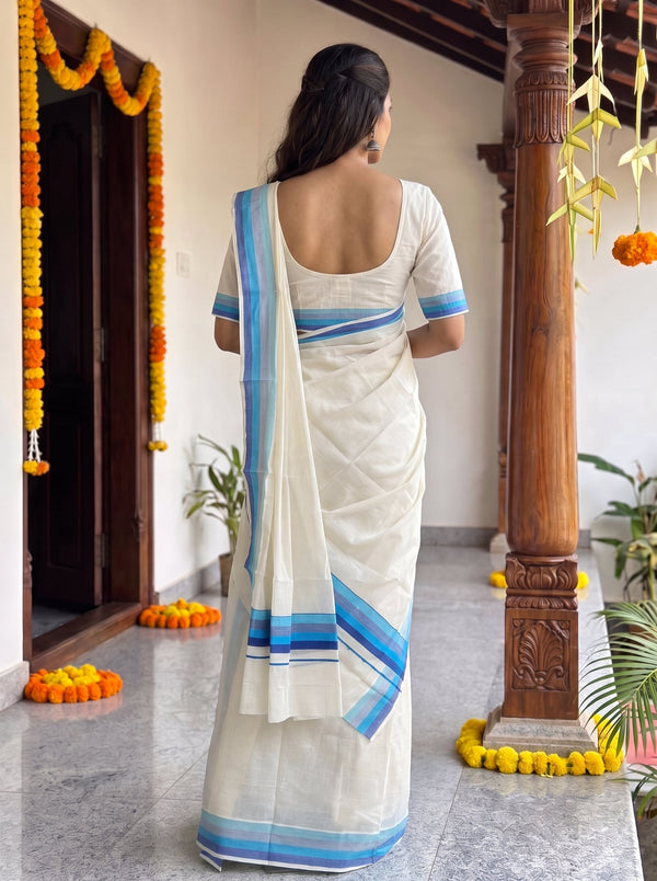 A woman viewed from behind wearing a white cotton saree with blue and light blue striped borders, standing on a tiled porch decorated with marigold garlands.