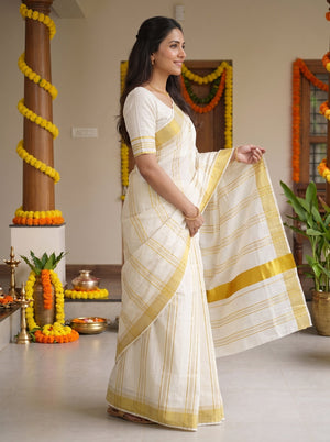 A woman wearing a white and yellow striped cotton saree stands in a decorated indoor setting, holding out the saree's pallu to display its pattern and border.