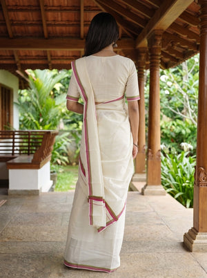 A woman viewed from behind wearing a cream-colored cotton saree with pink border accents, standing on a wooden porch with lush green garden foliage in the background.