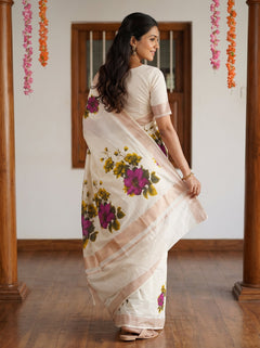 A woman wearing a cream-colored saree with a floral print in purple, green, and yellow, paired with a matching cream blouse, standing in a room with wooden pillars and a wooden floor.