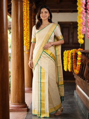 A woman modeling a cream-colored cotton saree with yellow and green borders, standing in a traditional corridor decorated with yellow and orange marigold garlands.