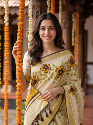 A woman wearing a beige and yellow floral saree with yellow and black border details, standing in a decorated courtyard with hanging marigold garlands.
