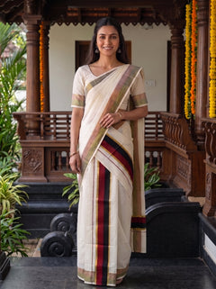 A woman wearing a traditional Indian saree stands in front of a wooden porch decorated with yellow flower garlands. The saree is off-white/cream with a striped border in red, green, and gold, paired with a matching cream blouse.