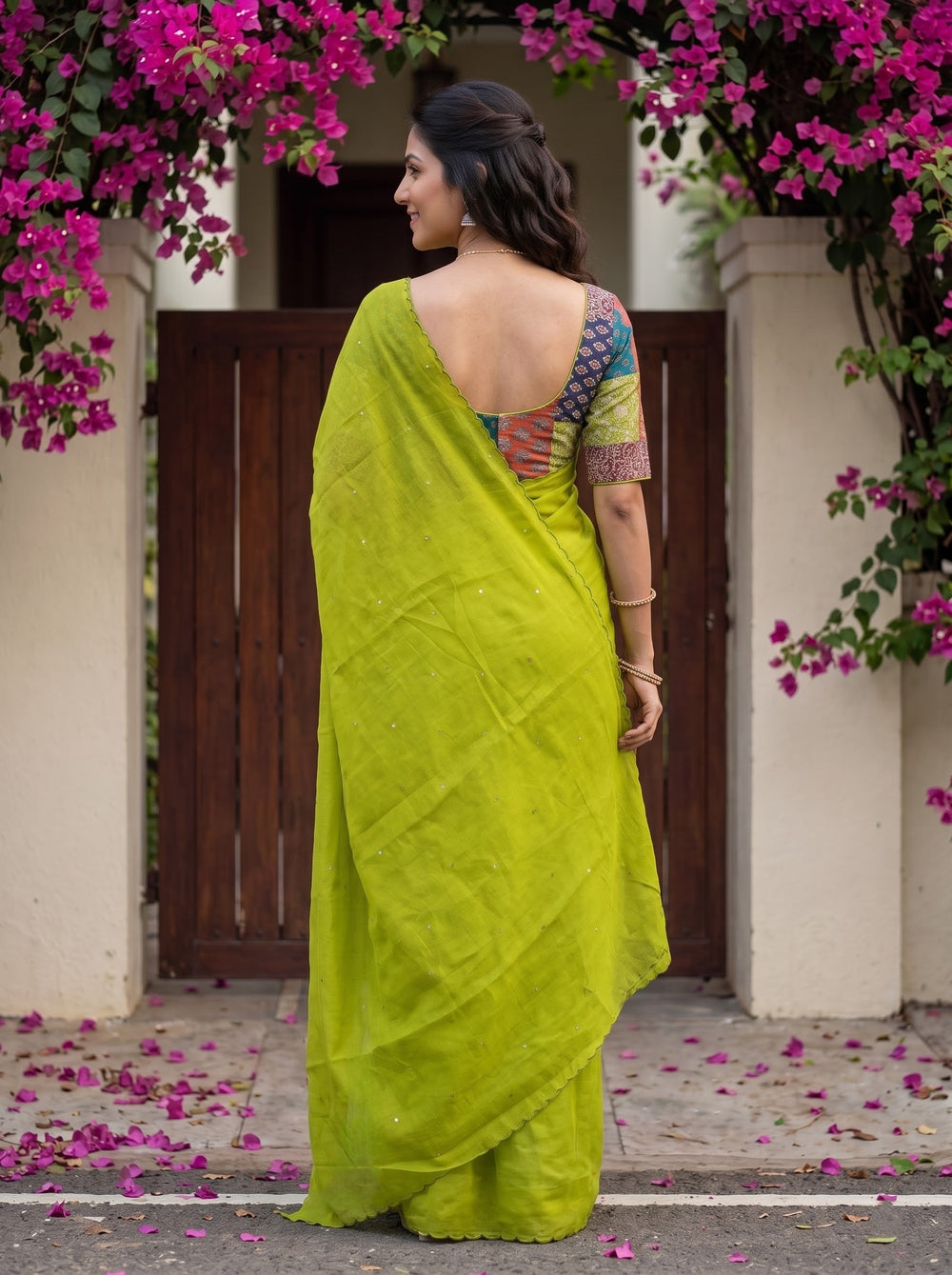 A woman wearing a bright lime green silk saree with a multicolored printed sleeve, standing in front of a rustic wooden door framed by pink bougainvillea flowers.