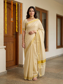 A woman wearing a cream-colored cotton saree with yellow border stripes, standing in front of a doorway decorated with orange marigold garlands.