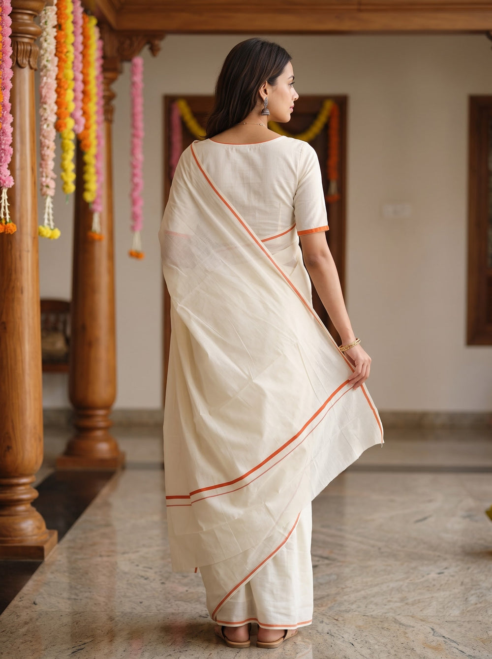 A woman wearing a cream-colored cotton saree with orange border stripes, standing in a decorated indoor space with wooden pillars and colorful garlands.