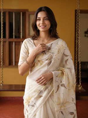A woman wearing a cream-colored sari with a beige and brown floral pattern, standing in a warm-toned indoor setting with wooden hanging swings in the background.