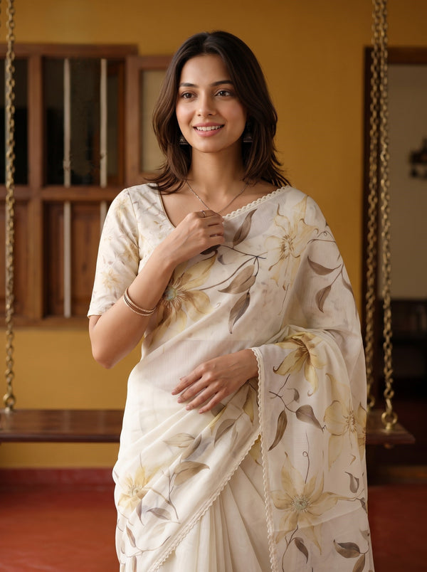 A woman wearing a cream-colored sari with a beige and brown floral pattern, standing in a warm-toned indoor setting with wooden hanging swings in the background.