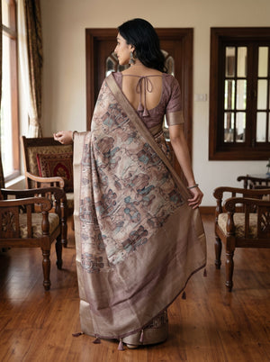 A woman stands in a room with wooden floors and chairs, wearing a draped saree with a muted mauve blouse and a patterned beige saree with blue and brown motifs.